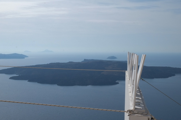 Photograph of a cinema sign from the side at a high elevation. The sign is made of individual letters screwed to a base. One letter is leaning backwards more than the others. Cables hold the sign in place on a ledge. In the background, the Santorini caldera islands are visible and disappearing in haze on the horizon. Santorini, Greece, 21 November 2019. Photo Cindy Kohtala