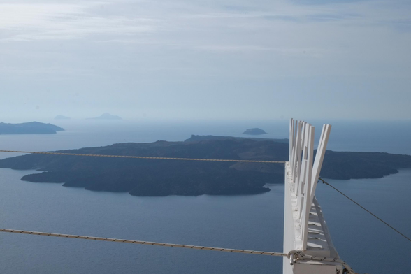 Photograph of a cinema sign from the side at a high elevation. The sign is made of individual letters screwed to a base. One letter is leaning backwards more than the others. Cables hold the sign in place on a ledge. In the background, the Santorini caldera islands are visible and disappearing in haze on the horizon. Santorini, Greece, 21 November 2019. Photo Cindy Kohtala 