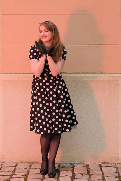 Une jeune femme aux cheveux long roux, posant devant un mur blanc, portant une robe noire à pois blanc, des collants noirs, des chaussures noires avec un petit nœud, des gants noirs et un serre tête noir avec des motifs blancs. Ses mains forment un V sous son menton et sa tête est tournée un peu vers sa droite, les yeux vers la caméra et un sourire enjoué 