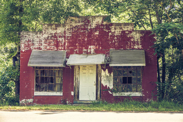 Abandoned single-story commercial building with severely deteriorating dark red paint exposing white or gray surfaces underneath. The symmetrical facade features a central white door with peeling paint, flanked by two large multi-pane windows with metal awnings above. The flat-roofed structure has a stepped parapet design creating three levels across the roofline. Dense green foliage and trees fill the background. Overgrown grass surrounds the building's foundation, and a paved road runs along the foreground. The building shows extensive weathering, paint loss, and general neglect. Taken in rural Georgia.