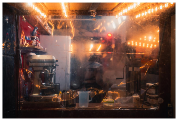 Seitlicher Blick durch eine krumpfelige Plastikfolie in einen Weihnachtsmarktstand. Hinter der Folie Küchengeräte, Lampen, eine Frau, Krimskrams.