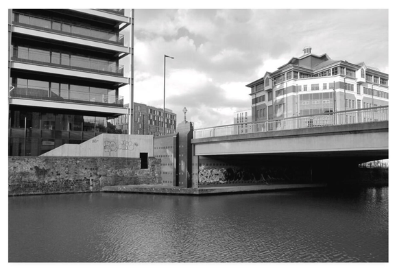 Black and white photograph of a modern cityscape with a river below and properties alongside the bridge above.  Graffiti is on the walls above and below the bridge.