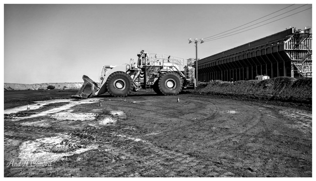 A wide view, black and white photograph signed by Kev Peirce. A massive front end wheel loader is positioned centrally on a heavily churned, muddy, and dark earth surface.

Its large bucket rests on the ground to the left. To the right, a long, dark industrial structure, possibly a conveyor system or processing plant, dominates the background.

A utility pole stands near the structure. The composition emphasises the heavy, industrial nature of the site and the contrast between the machine and the facility.