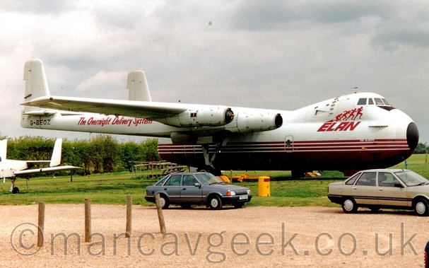 Side view of a high-winged, 4 propellor-engined cargo plane parked facing to the right on grass as part of an external museum exhibit.
The plane is largely white, with a maroon lower half, and red "Elan" titles under red images of an athlete starting to run out of starting blocks on the upper forward fuselage, under the cockpit which is in a bump on the forward fuselage.
The plane's fuselage is quite short, finishing not far behind the wings.
2 engines are mounted on each wing, the inner engine pod holding both the main undercarriage, and a long tail boom, which extends rearwards to the twin tails which have  a large horizontal stabiliser mounted between them.
A pair of cars, one grey, the other brown, are parked on a sandy gravel track in the foreground, with trees in the background lining the Aero park's perimeter, under cloudy grey sky.
A much smaller white plane is trying to squeeze into frame on the left.