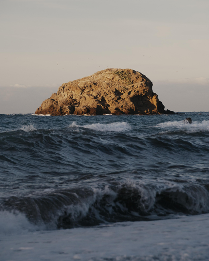 "Sunlit rocky island surrounded by crashing ocean waves under a clear late-afternoon sky."