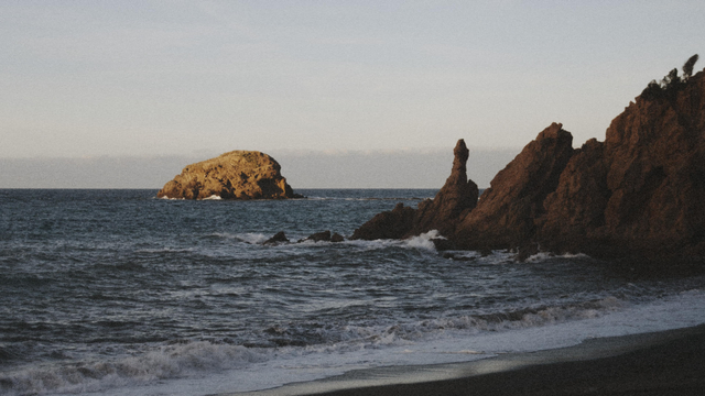 Sunlit offshore rock formation beyond dark sand beach and crashing waves under a clear sky."  
Emphasizes the contrast between light and shadow, motion and stillness.