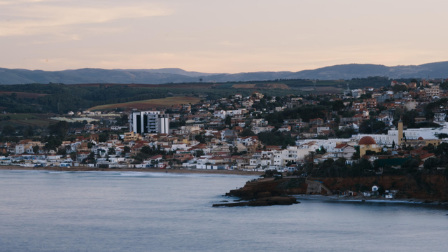 "Densely built coastal town climbing hillside, with pastel sky and calm sea at sunset."