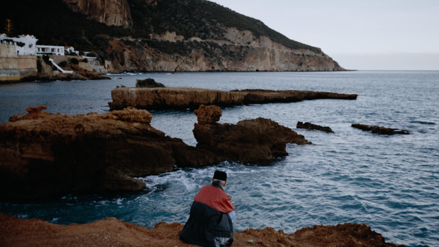 "Solitary figure in red and black sits on cliffside overlooking deep blue sea and rugged coastline under overcast sky."