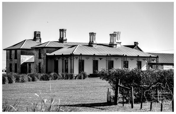 A striking black and white photograph of Highfield House, showcasing its impressive row of chimney stacks and veranda. 

The historic colonial house is set against a bright sky, framed by foreground grasses and a small wooden fence.