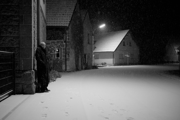 Dorf im dunklen. Straße entlang fotografiert. Die Straße biegt nach rechts ab. Links Häuser. Dunkel Beleuchtung. Ich stehe unter einer Lampe. an die Lampe gelehnt. Stehe seitlich zur Kamera. Schaue in die Kamera. Mantel an und Jeans. Sneaker. Glatze. Brille. Straße Schnee bedeckt und es schneit.