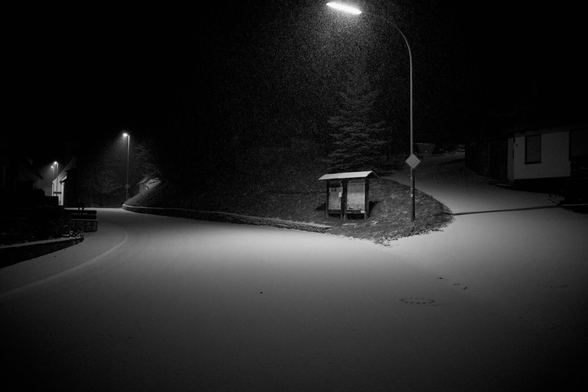 eine Straße biegt nach links ab. Die andere den Berg hoch nach rechts. Dorflampen entlang der Straße. Zwischen den Straßen sind Schilter. Vereinzelt Häuser. Dunkel. Beleuchtung.Straße Schnee bedeckt und es schneit.