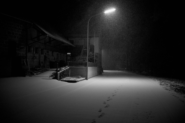 Straße entlang fotografiert. Links ein Bauernhof. und ne Dorflampe. Rechts Wiese und dunkel. Beleuchtung Lampe. Es schneit und die Sachen sind Schnee bedeckt.