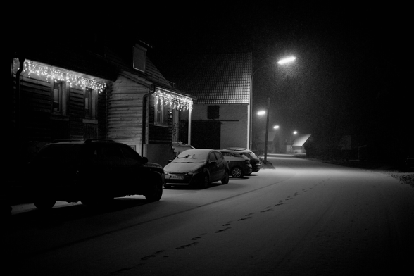 Ein Haus mit Lichterketten am Haus. Straße entlang fotografiert. Autos stehen Links. Eine Halle neben den haus. Die Straße führt in die ferne. Entlang der Straße Häuser und Dorflampen die Leuchten. Autos und Straße Schnee bedeckt. Es schneit.