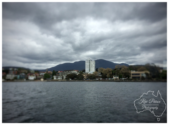 A photograph showing the approach to Hobart, Tasmania, with a focus on a tall, white building in the center. 
The foreground is a dark, slightly blurred body of water (the Derwent River).

The mid ground features the shoreline with low rise buildings and trees, and the background is dominated by the dark, majestic outline of Mount Wellington under a dramatic, cloudy sky.

The image has a tilt-shift effect, blurring the top and bottom.