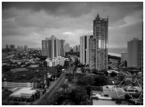 A high angle, black and white photograph of the Surfers Paradise skyline, featuring several high rise residential towers and hotels under a dramatic, cloudy sky.

The ocean is visible on the far right. A prominent central tower dominates the frame, with a main road running through the foreground.

The image has a selective focus or tilt shift effect, blurring the lower and top edges.