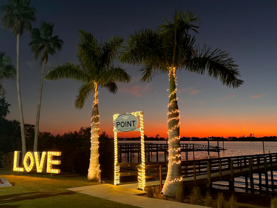 Photograph by Fenichel

A view of a Bay at sunset, a vivid red at the horizon line, and deep blue above.
In the middle/center are two palm trees wrapped with yellow Christmas lights, and between the trees, a rectangular 'arch', also strung in small lights, and with a sign top/center saying 'POINT'.

In the bottom 1/3 (foreground), left to right is a big sign on the ground (again, in yellow light) big box letters forming the word, "LOVE". To the right, the bottom of the trees and 'archway" Past the door/arch (bottom right quarter) is a dock extending out over the water. 