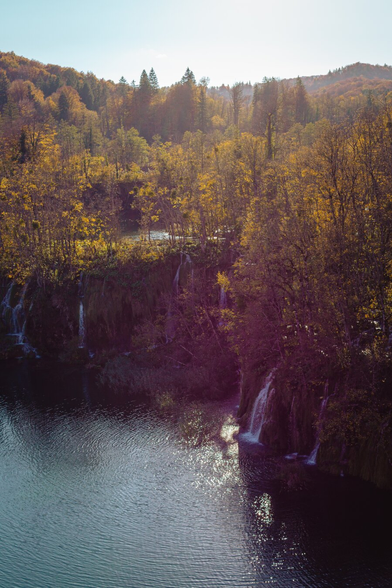 A lake surrounded by forest and fed by waterfalls, the sun hangs low, illuminating the scene.