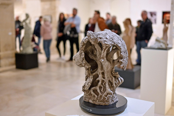 Photo de l'exposition d'art avec une sculpture au premier plan : une tête d'ours de 3/4 dos ouverte sur l'arrière et porté par un arbre sur les racines duquel une jeune femme est assise en train de lire. En arrière plan, une quinzaine de personnes debout sont floues. ainsi que d'autres sculptures sur des socles et photographies sur les murs