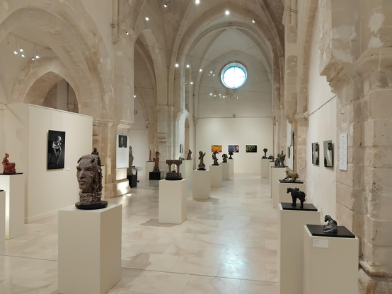 Photo en plan large d'une exposition d'art (sculptures et photographies) dans une ancienne église en pierres blondes. On y distingue une vingtaine de sculptures sur des socles blancs et une dizaine de photographies sur les murs.