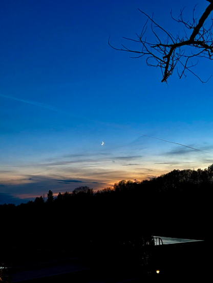 A serene twilight sky features a faint crescent moon amidst blue and orange hues, with silhouettes of trees and a roofline in the foreground.