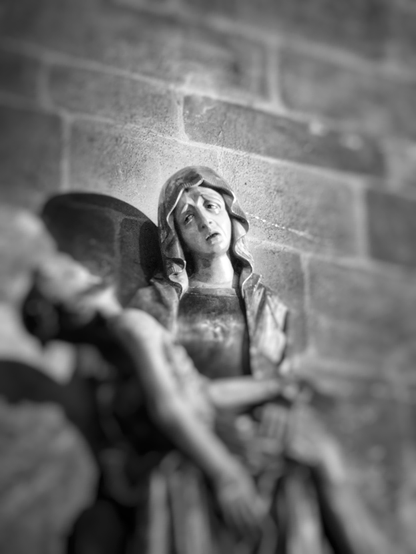 Schwarzweißaufnahme einer Frauenfigur mit verzweifeltem, schmerzerfülltem Gesicht. Der Kopf ist leicht zur linken Bildseite geneigt. Sie trägt ein Tuch auf dem Kopf, das bis über die Schultern fällt. Ihr Mund ist geöffnet und ihr Blick ist zur linken Bildseite gerichtet. Alles andere auf dem Bild, wie die gemauerte Wand hinter der Figur oder die männliche Figur, die sie in den Armen hält und auf ihrem Schoß liegt, ist unscharf. 