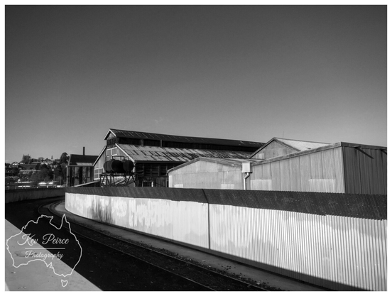 Black and white photograph of the Launceston Old Railway Workshops. The foreground is dominated by a sharply curving railway track running parallel to a long, corrugated metal fence that reflects the bright light.  Behind the fence, several large, old railway maintenance sheds and industrial buildings with corrugated iron roofs rise up against a clear, bright sky.