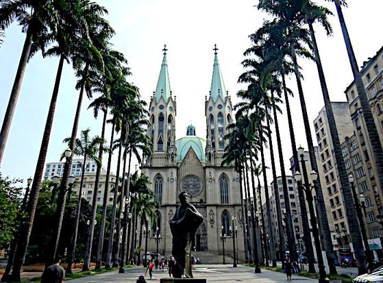 Foto van standbeeld tussen twee rijen palmen en op de achtergrond de cathedraal van São Paulo