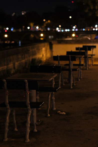 Photo nocturne de tables avec plateau d’echec intégré, toutes vides, légèrement éclairées par une lumière orange. Les lumières de la ville floue en fond