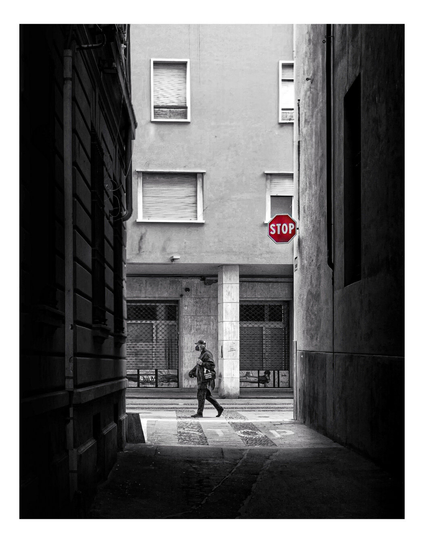 Black and white photograph looking down narrow alleyway. A lone man is at the end of it with a backpack and a hat walks past a red stop sign on the right. The alley is enclosed by tall buildings with textured walls.  The ground is paved with concrete. The man is dressed in dark clothing. The red stop sign is the only color in the image.