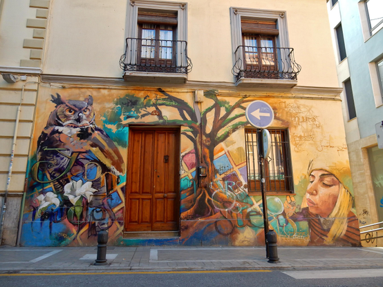The front of an old building with ornate windows and a large wood door. The front is covered with a beautiful picture of an owl, the trunk and limbs of a tree, and someone blowing bubbles.