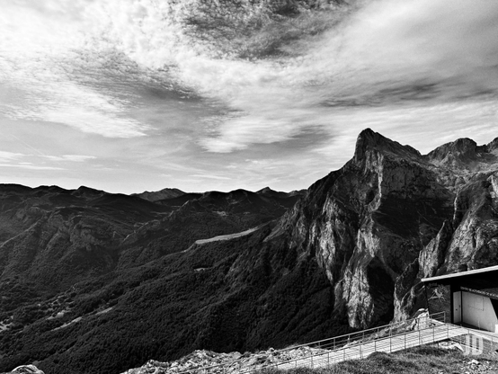 Fotografía en blanco y negro de un vasto paisaje montañoso. El cielo está cubierto por nubes estratificadas y dispersas. Se aprecian múltiples cadenas montañosas; las laderas y montañas a la izquierda están densamente cubiertas de bosque, mientras que la montaña de la derecha es un pico rocoso, escarpado e imponente, con fuertes contrastes de luz y sombra. En primer plano, a la derecha, una moderna edificación con techo inclinado y una entrada clara, identificada como 'CENTRO DE ACTIVIDADES DE MONTAÑA', se integra en la ladera. Una pasarela con barandillas metálicas conduce hacia su entrada sobre terreno herboso. La imagen en blanco y negro enfatiza la profundidad y las texturas rugosas del entorno.