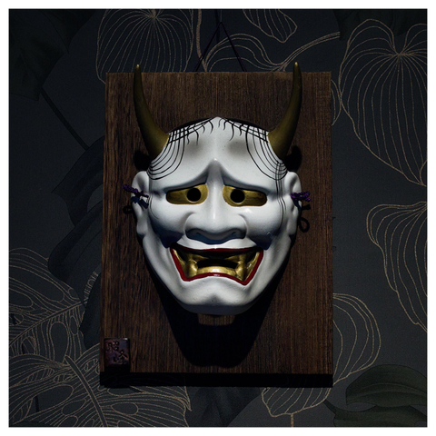 En fyrkantig bild på en vit mask med horn. Masken har röda läppar och stora tänder och ser skräckinjagande ut och sitter på en träbit, upphängd på en vägg med mörk, mönstrad tapet.