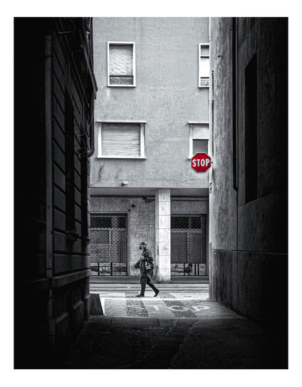 Black and white photograph looking down narrow alleyway. A lone man is at the end of it with a backpack and a hat walks past a red stop sign on the right. The alley is enclosed by tall buildings with textured walls.  The ground is paved with concrete. The man is dressed in dark clothing. The red stop sign is the only color in the image