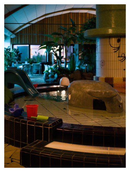 En stående bild på barnavdelningen inne på ett badhus. Diverse leksaker vid ett mycket grund bassäng. En stor sten är målad med val-ansikte, en pelare har målat ansikte. 