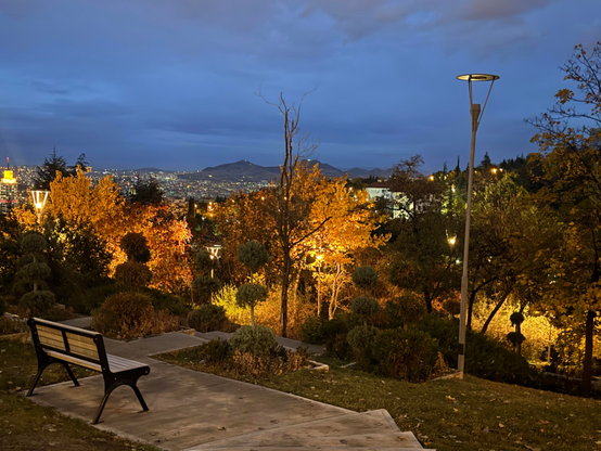 A park scene at dusk with a bench overlooking Ankara city full of lights. Trees with autumn leaves glow under warm lamplight, and distant hills are visible under a deep blue sky.