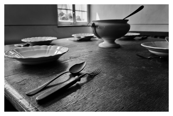 The image is a black-and-white photograph of a rustic dining table. In the foreground is a place setting consisting of a white plate with a scalloped edge, a spoon, and a fork, which lie parallel to each other next to the plate on the wooden surface of the table. A piece of the spoon appears to be broken off, and the prongs of the fork are heavily worn and slightly bent. There are four additional empty plates with cutlery on the table, arranged in a circle around a large, ornate serving bowl with a ladle. The table appears worn, with visible grain and texture. In the background, a window with multiple panes lets in soft light that illuminates the scene and casts gentle shadows. The overall atmosphere is calm and old-fashioned, suggesting a historical or vintage setting.