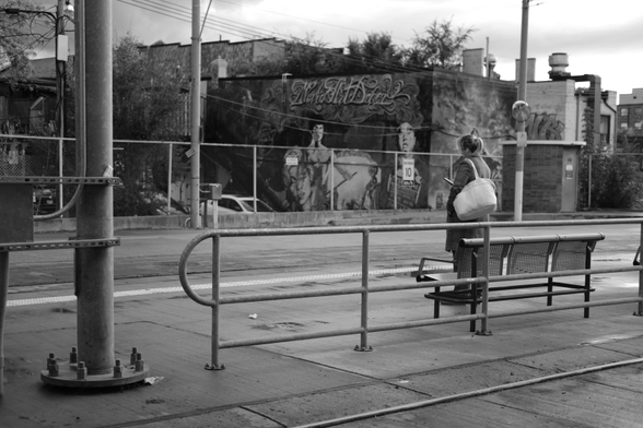 The outdoor streetcar platform of the station. At extreme left, a utility pole runs from top to bottom of the image. Just right of centre, a single woman stands in front of a bench, waiting for the 505 Dundas car. In the background, beyond the chain link fence that boundaries the station, are murals painted on the buildgings beyond. 