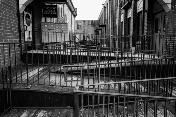 A black and white photo of a series of wooden ramps with hand rails leading up to an entryway between buildings and a mail kiosk.