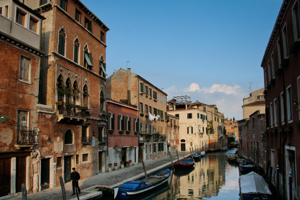 A canal in Venice with moored boats, historic buildings, and a person walking along the waterfront.