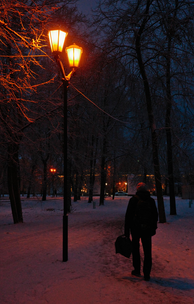 Late evening. Park. The lantern is glowing orange. A man stands under a street lamp. There is snow on the ground.