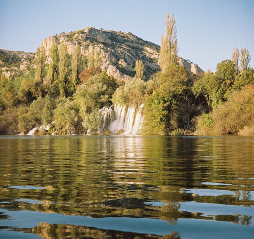 The edge of the mountain from which the waterfall flows into the lake. The rocks are covered with greenery, many trees are golden. The scene is reflected in the water.