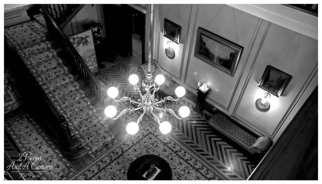 An overhead, high angle, black and white photograph looking down into the main hall and stairwell of Martindale Hall. 

The image is dominated by a brightly lit, multi bulb chandelier hanging centrally. To the left, a grand, dark wooden staircase covered in a patterned carpet descends.

Below the chandelier, the floor features a striking zigzag parquet pattern, partially covered by a large, ornate area rug.

The walls, panelled in wainscoting, display framed paintings and wall sconces. The photograph captures the symmetrical grandeur of the entrance space. The photograph is signed Kev Peirce.
