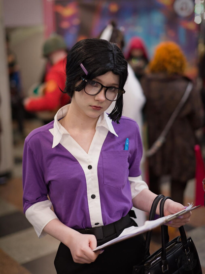 A person stands facing slightly to the right. They wear a short-sleeved, two-toned shirt: purple on the front and sleeves, white on the collar and chest. The shirt has a black button down the front, a pocket holding a blue pen, and white inner sleeves. They also wear black skirt, a black belt, a purple hair clip in their dark hair, and black-framed glasses. In one hand, they hold a clipboard; the other hand grips a black handbag. The background is blurred, showing other people in varied outfits, suggesting a public event like a convention.