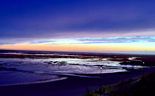 Ein Sonnenuntergang in Wittdün auf Amrum bei dem sich zwischen zwei Wolkenbanken ein Streifen Orangen Abendlichts zeigt, während auf dem dunklen Kniepsand-Strand Wasserflächen silbern und blau schimmern.
