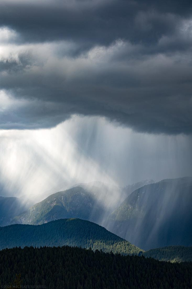 A photograph of dark clouds over a series of mountain peaks, with a downpour of rain hitting them, and side light from the sun shining beams directly onto the torrents of rain.
