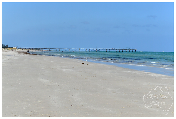 A tranquil colour photograph capturing the wide, sandy beach and the historic Semaphore Pier in Adelaide, South Australia.

The long, horizontal pier extends over the turquoise water of Gulf St Vincent into the distance under a vast, cloudless light blue sky.

The foreground is dominated by the pale, sun-drenched sand, with the water gently lapping the shore.