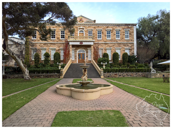 The impressive, stately stone facade of Chateau Yaldara winery in the Barossa Valley, featuring two stories of arched windows, a central entrance accessed by a wide stone staircase, and a decorative fountain in the foreground leading up to the main building via a brick pathway. The building is surrounded by well manicured green lawns and topiaries.