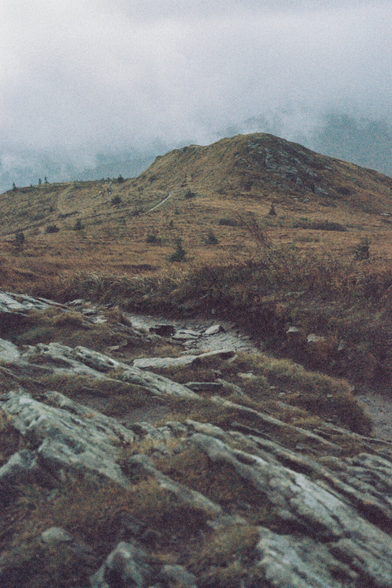 A muddy path leading through heathland and dry grass, with mist-covered mountains and breaking clouds in the background.
