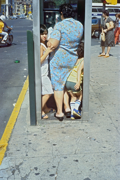 The photo captures a candid moment of a woman and two children squeezed into a public phone booth on a New York City street. 