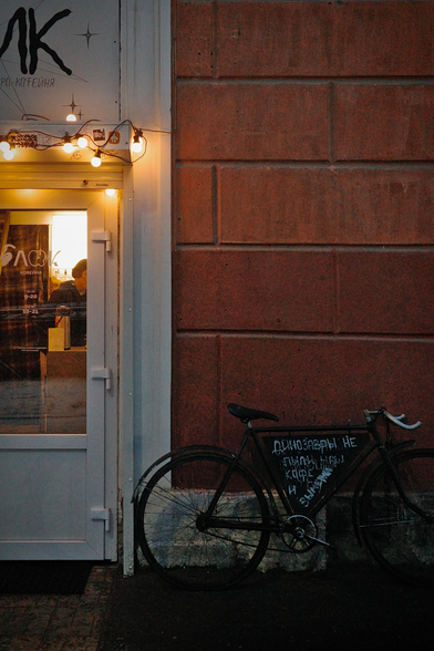 In the photo, a black bike stands against a red wall next to the door of the coffee shop. Late evening. The bike is illuminated by the warm lights of the coffee shop. There is an inscription on it: "Dinosaurs did not drink our coffee and became extinct."